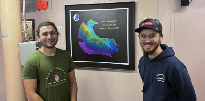 Kaan Cav and Lloyd Pikok stand in front of a wall-mounted poster of the Healy Seamount bathymetric image.