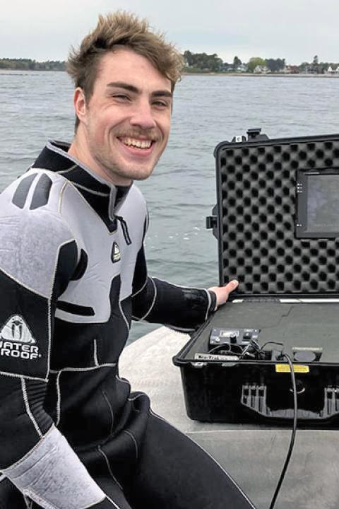 Grant Milne in wetsuit sitting on open boat with equipment case.